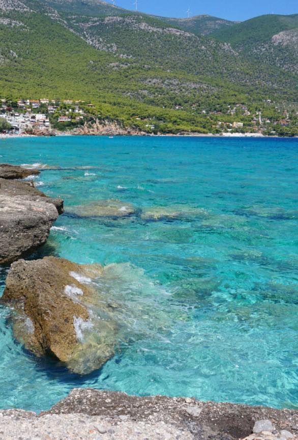 Clear turquoise water along a rocky shoreline near Porto Germeno, with the village in the distance